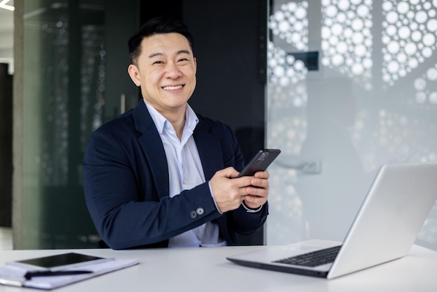 Happy asian man using phone in office businessman smiling and looking at camera typing message and