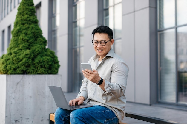 Happy asian middle aged businessman texting on cellphone and using laptop chatting with client outdoors