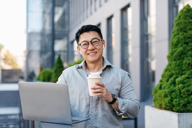 Happy asian middle aged businessman working on laptop and drinking coffee outdoors near office building