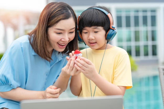 Happy Asian mother gives a toy to her son while boy using headphones with laptop at home