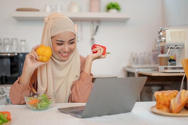 A happy Asian Muslim woman in a hijab is working on her laptop at a dining table in the kitchen