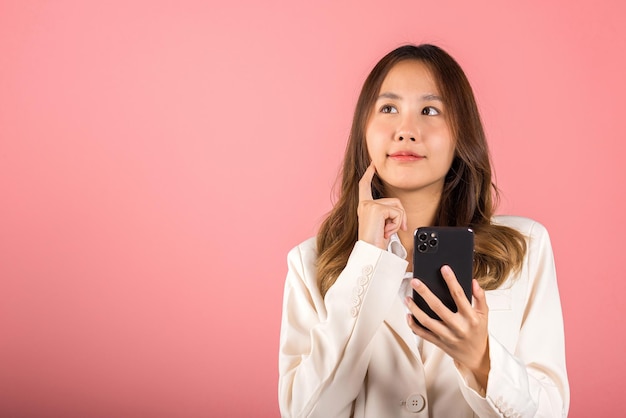 Happy Asian portrait beautiful cute young woman excited smiling holding mobile phone and think idea finger touch face, studio shot isolated on pink background, female using smartphone making gesture