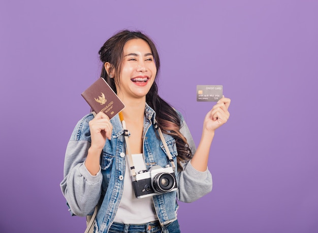 Happy Asian portrait beautiful young woman excited smile in summer holding passport, credit card and vintage photo camera, Thai female ready travel trip looking side away isolated on purple background