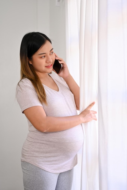 Happy Asian pregnant woman looking out the window and talking on the phone with her husband