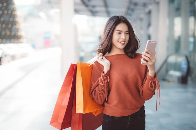 Happy Asian pretty girl holding shopping bags while looking at smart phone 