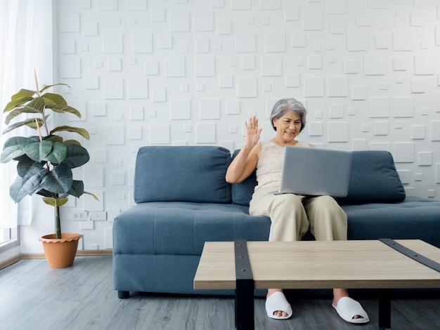 Happy Asian senior casual woman sitting on the sofa waving greeting at laptop computer screen in white room Elderly female meet with family by video call at home Older people with technology