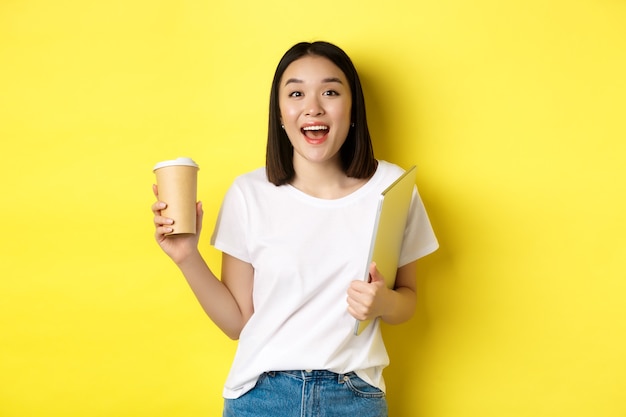 Happy asian woman drinking coffee and holding laptop, saying hi to you, standing over yellow background.