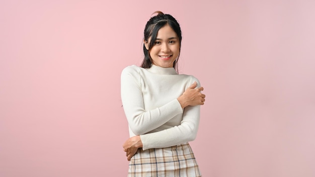 Happy Asian woman embracing herself with love and kindness isolated on pink background