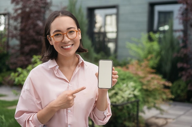 Happy asian woman holding mobile phone shopping online pointing finger at display. Mockup