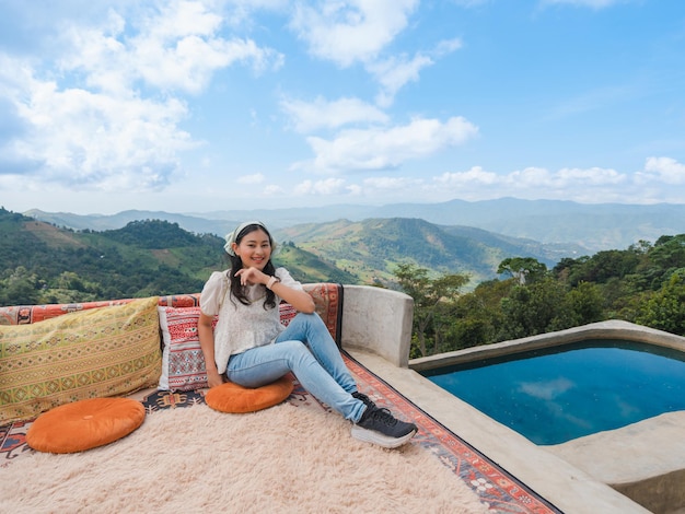 Happy Asian woman sit and relax at balcony with mountain view with cloudy blue sky at Doi Chang, travel attraction in Chiang Rai, Thailand