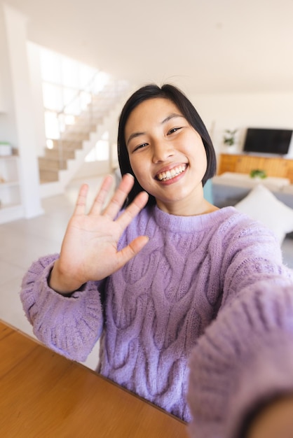 Photo happy asian woman sitting at table and having video call, waving to camera. spending quality time at home, domestic life and lifestyle concept.