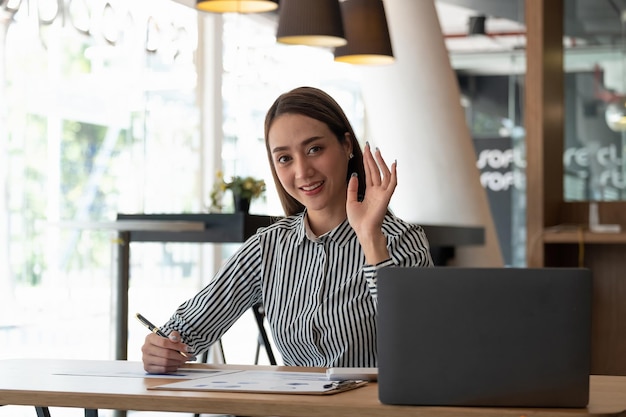 Happy asian woman smiling and waving hand at laptop while speaking or chatting on video call in office online conference webinar concept