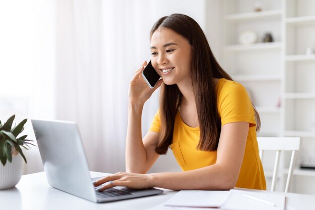 Happy asian woman talking on cellphone while working on laptop at home