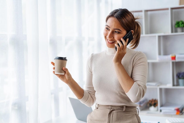 Happy asian woman talking phone while holding coffee cup and standing at desk in office workplace