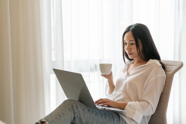 Happy Asian woman using laptop while holding a cup of coffee at home Smiling girl working on computer enjoying internet shopping or chatting in social networks Technology and lifestyle concept