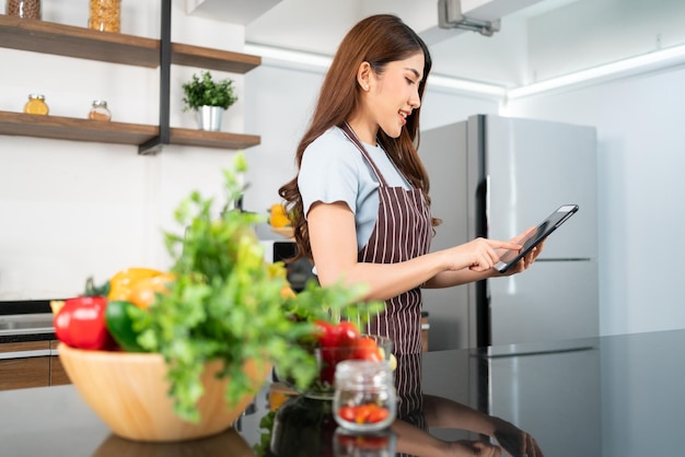 Happy Asian woman wearing an apron prepares vegetables for healthy food by searching recipes online on a digital tablet in home kitchen watching videos of ingredients Modern cooking technology