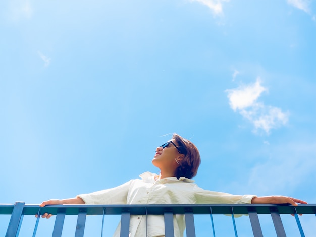 Happy Asian woman wearing white shirt and sunglasses standing on the balcony on blue sky background.