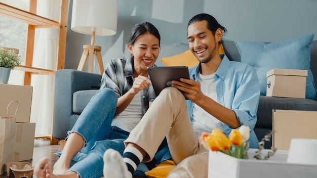 Happy asian young couple man and woman use tablet for online shopping furniture to decorate house with carton packages in new house.