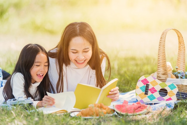 Happy Asian young family mother and child little girl having fun and enjoying outdoor laying on picnic blanket reading book at summer garden spring park, Family relaxation concept