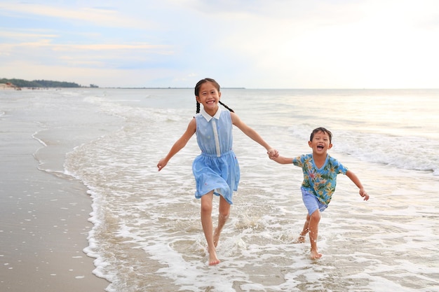 Happy Asian young sister and little brother having fun running together on tropical sand beach at sunrise Happy family boy and girl enjoy in summer holiday