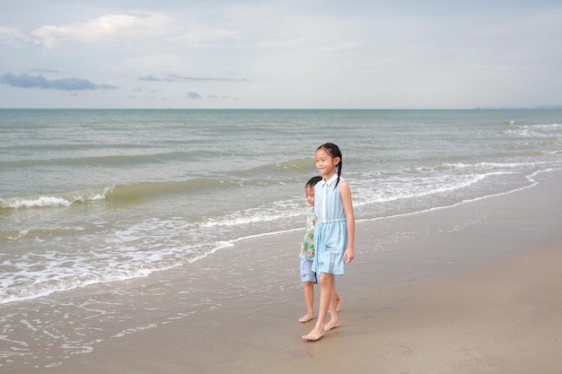 Happy Asian young sister and little brother walking together on tropical sand beach at sunrise Happy family boy and girl enjoy in summer holiday