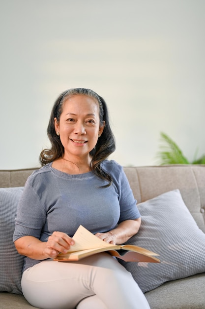 Happy Asianaged woman sits on a sofa in her living room with a book in her hands