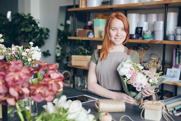 Happy attractive redhead young florist in apron holding beautiful bouquet while working in own shop