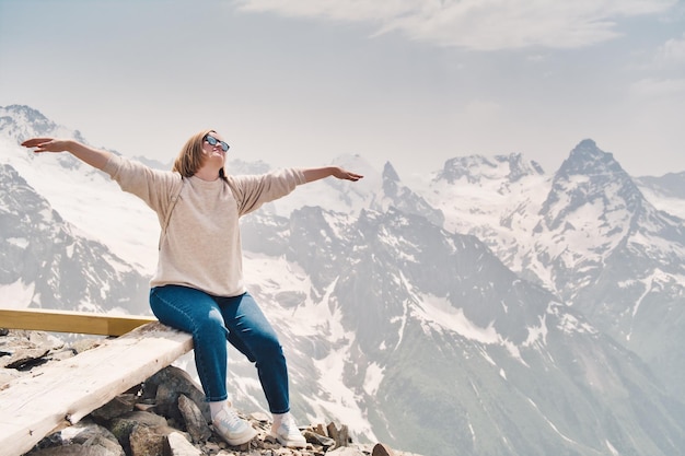 Happy attractive woman in sunglasses sits on a rock with raised hands. Happy tourist in mountains