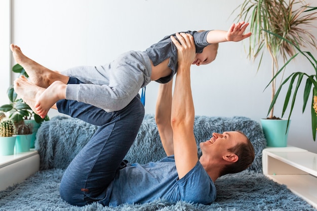 Happy baby boy flying in father's arms looking, loving family dad holding lifting cute little child son playing plane, bonding, having fun on couch