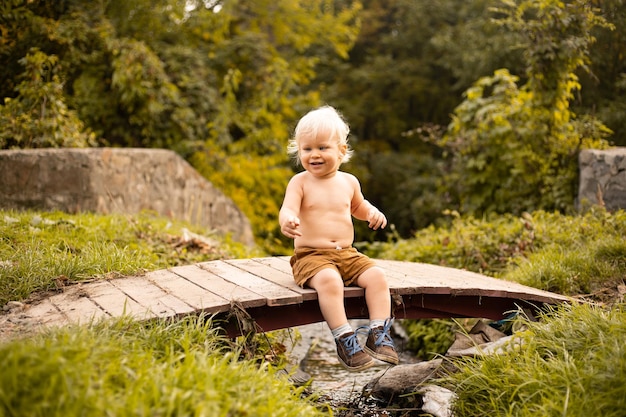 Happy baby boy sitting on bridge over the stream and smiling in autumn or summer park with golden trees