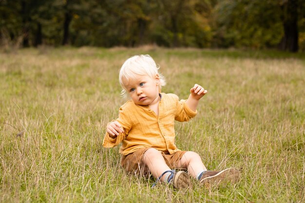 Happy baby boy sitting in meadow and smiling in warm autumn or summer park with golden trees