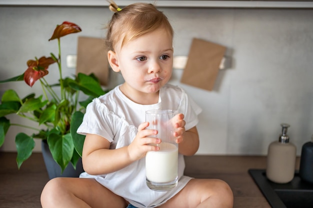 Happy baby girl sitting at the table in the kitchen and drinking milk. High quality photo