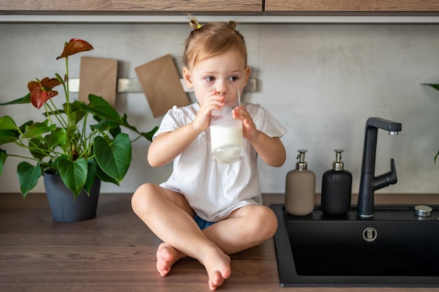 Happy baby girl sitting at the table in the kitchen and drinking milk