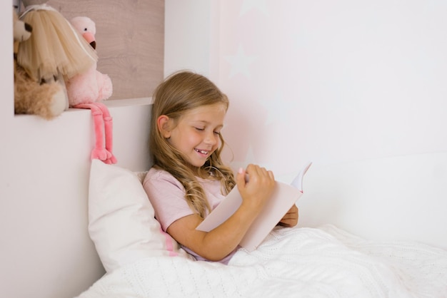 Happy baby girl with long hair reading a book on a bed with pillows