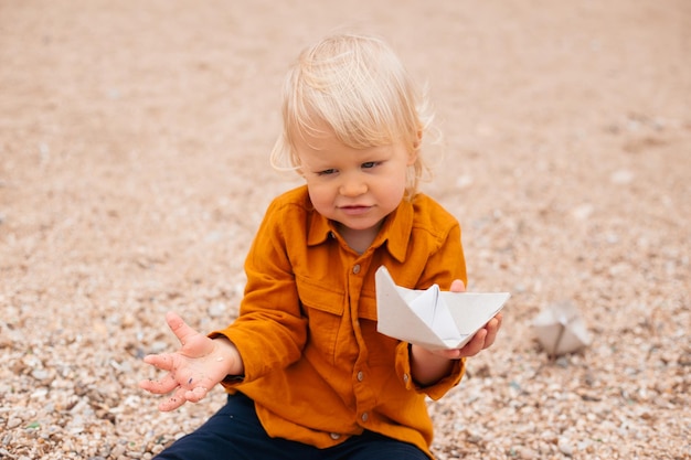 Happy baby playing with paper boat in the beach in autumn time