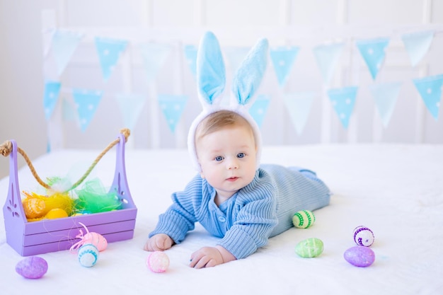 A happy baby with Easter bunny ears colorful Easter eggs and a basket of eggs is lying at home on a bed on white pastel linen Easter Greeting Card