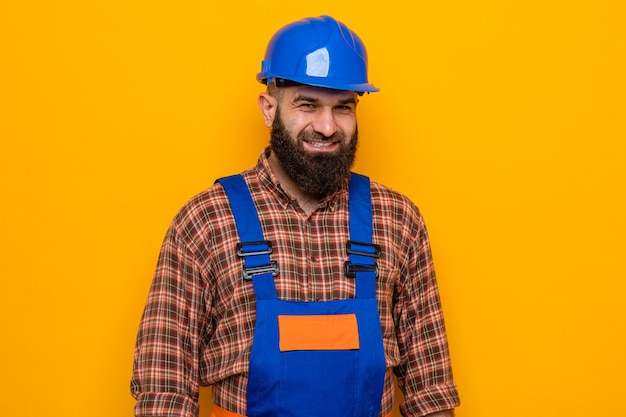 Happy bearded builder man in construction uniform and safety helmet looking at camera smiling cheerfully standing over orange background