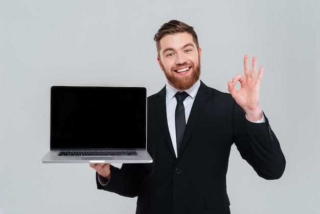 Happy bearded business man showing blank laptop screen and showing ok sign. Isolated gray background