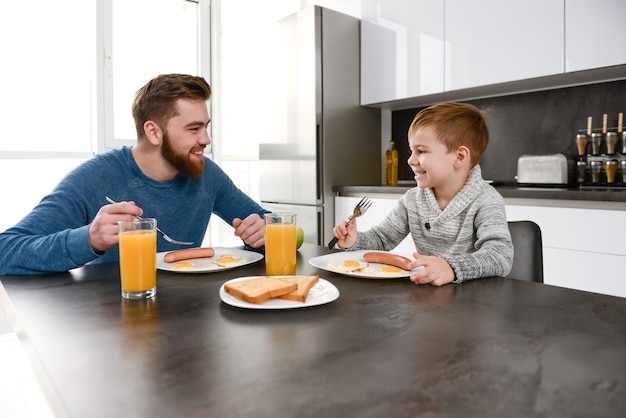 happy bearded father dressed in blue sweater eating at kitchen with his little son