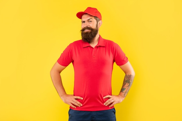 Happy bearded man in casual red cap and tshirt standing with tattooed arms akimbo yellow background man