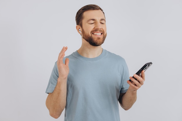 Happy bearded man dancing with earphones listening music in headphones holding mobile phone and smiling pleased standing over white background