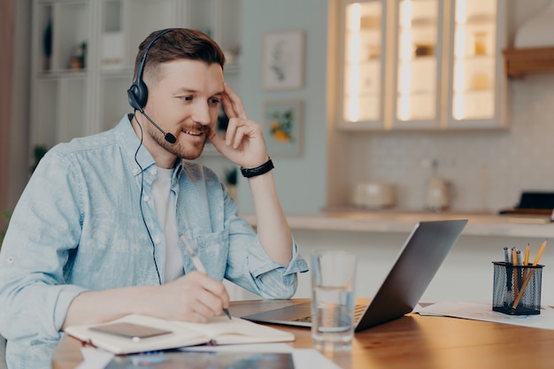 Happy bearded man with headset with microphone looking at laptop screen and making notes during distance education, sitting at his apartment while working online. Elearning and freelance concept