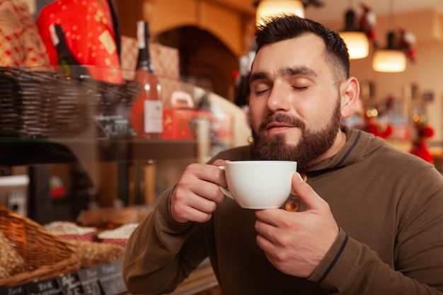 Happy bearded young man enjoying smell of fresh coffee, having breakfast at the cafe, copy space