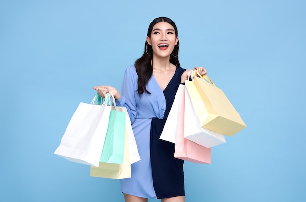 Happy beautiful Asian shopaholic woman wearing blue dress and holding shopping bags isolated on blue wall.