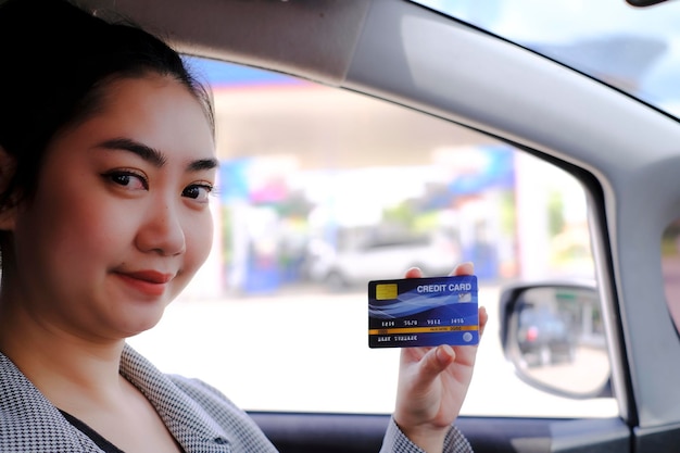 Happy beautiful Asian woman sitting inside her car showing credit card payment at a gas station