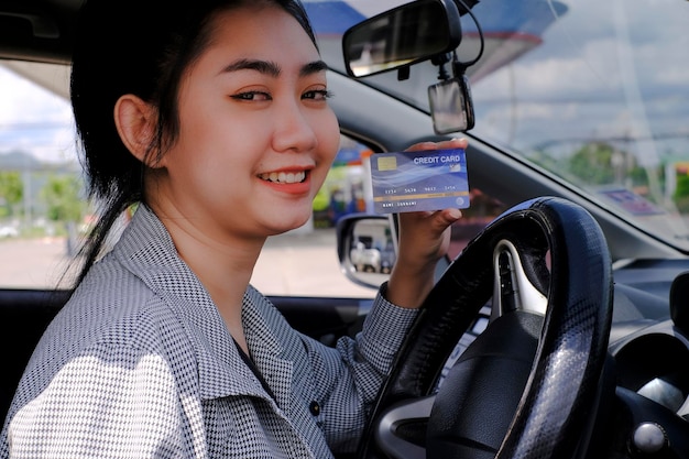 Happy beautiful Asian woman sitting inside her car showing credit card payment at a gas station