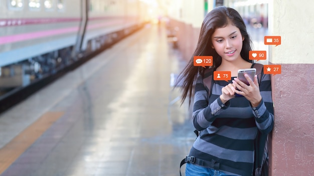 Happy and beautiful asian woman standing in train station use mobile phone with social media interaction, notification icons, e-mail, message, like, love, comment on device.  Lifestyle with technology