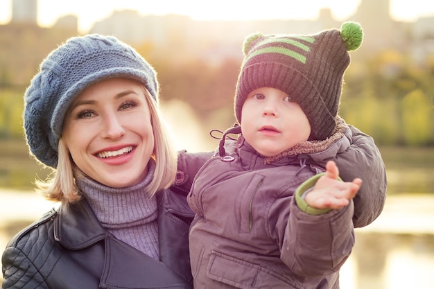 Happy and beautiful blonde woman in hat kissing baby son in a warm jacket holding him in her arms in the autumn park