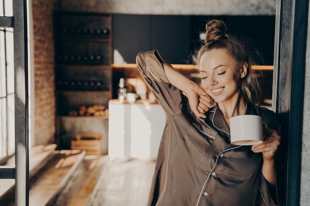 Happy beautiful brunette female stratching with cup of coffee in her hand while standing in kitchen