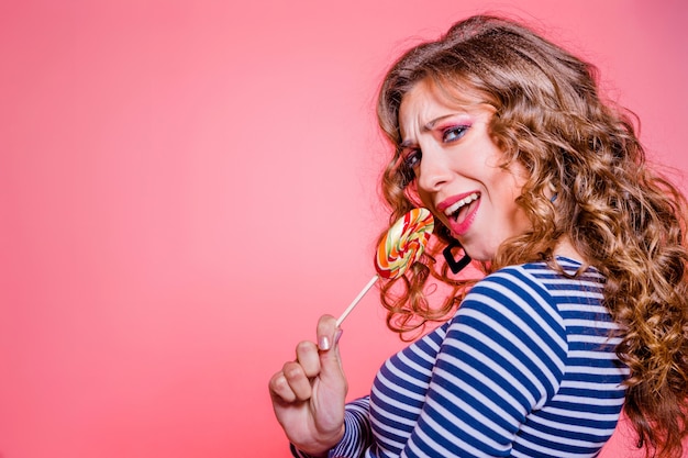 Happy beautiful brunette girl with red makeup, curly hair and a blue striped sweater, posing against a red background with a colored lollipop in her hands. Girl pretending to sing. Horizontal photo